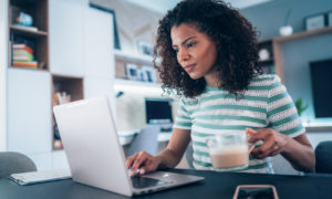 Mulher tomando café em frente ao notebook. Imagem simboliza que a profissional está aplicando boas práticas no modelo home office.