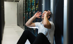 Homem com expressão de preocupado sentado perto dos servidores. Imagem simboliza problemas vindos de uma infraestrutura de TI inadequada.