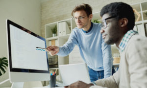 Dois profissionais conversando em frente ao notebook. Imagem simboliza que eles são desenvolvedores iniciantes no dia a dia de trabalho.