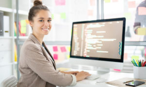 Mulher sorrindo em frente ao computador, em escritório. Imagem simboliza que ela está trabalhando com o no-code e low-code.