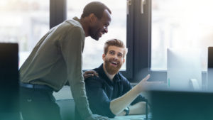 Dois homens conversando em frente ao computador. Imagem simboliza que são profissionais de TI aplicando tecnologias em pequenos e médios negócios.