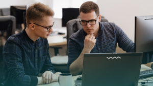 Dois profissionais conversando em frente ao computador. Imagem simboliza que são programadores falando sobre o core business da empresa.